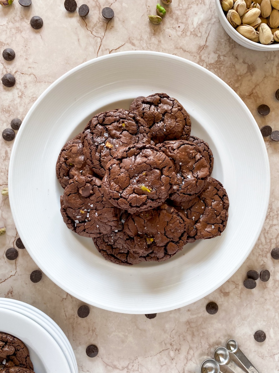 Crackle-topped brownie cookies sit on a white plate on a surface sprinkled with chocolate chips