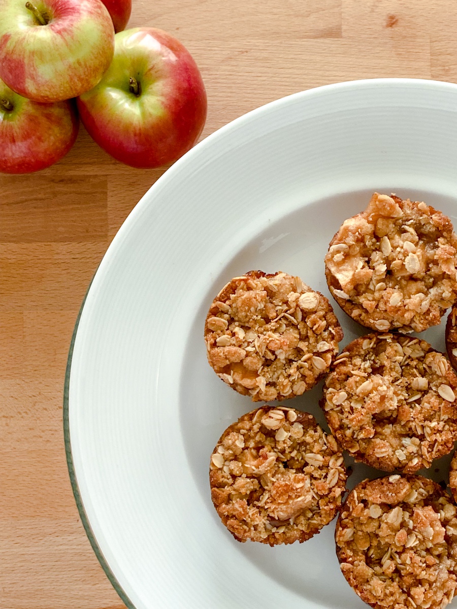 Mini pies topped with oatmeal crumble arranged on a white plate next to apples on a wooden surface.