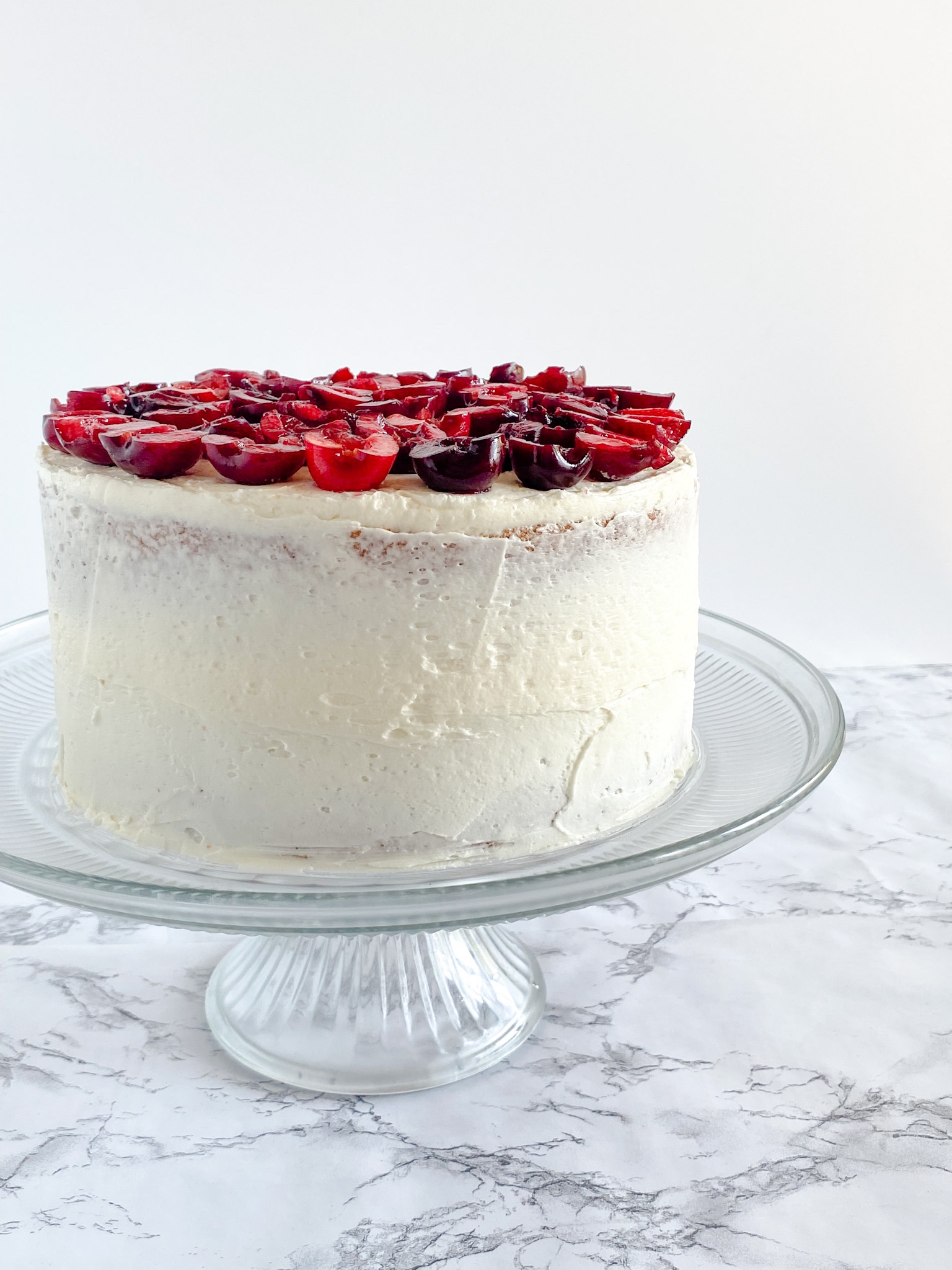 A lightly-frosted white cake topped with fresh cherries on a glass cake stand on a marble surface