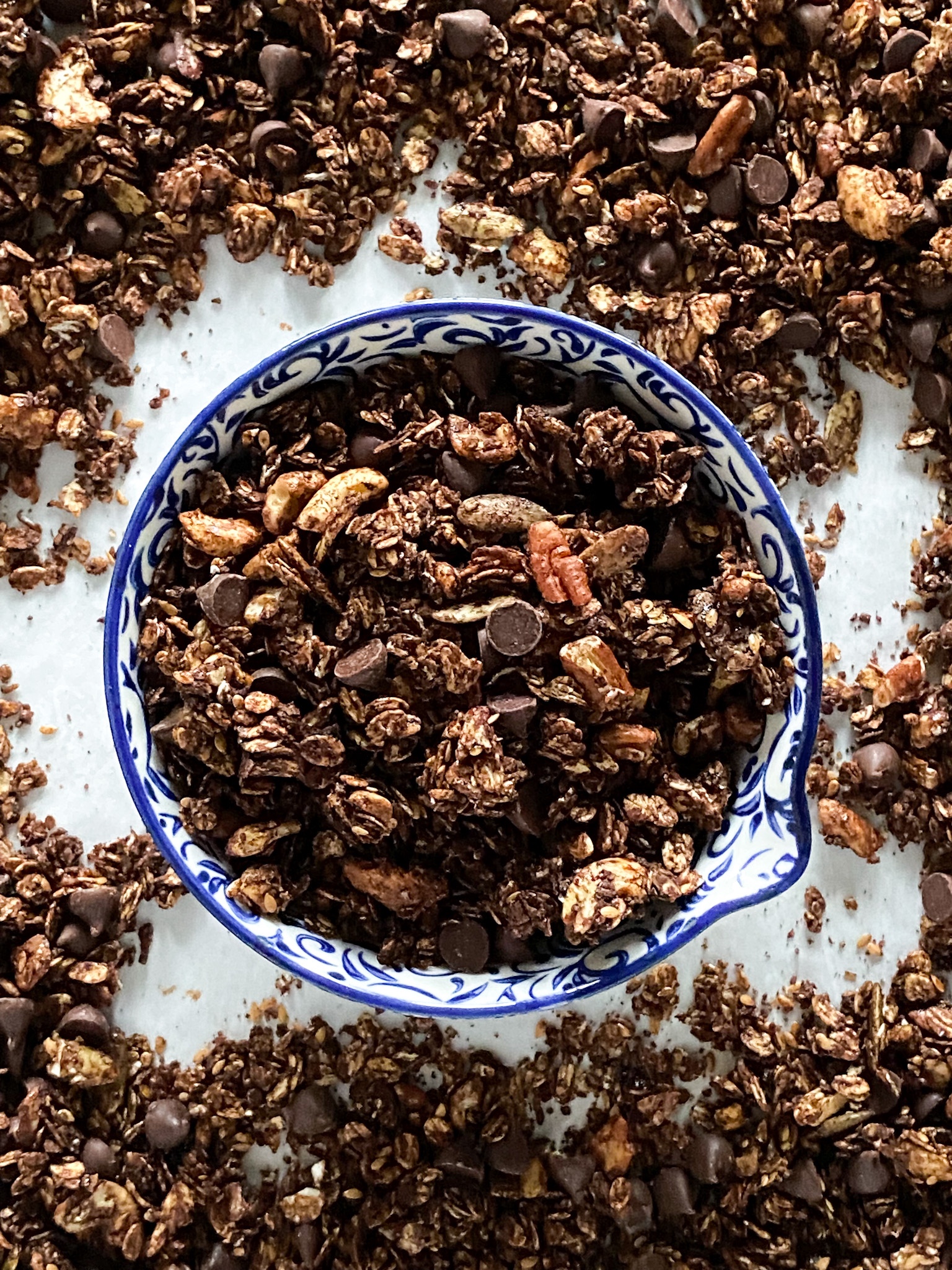 A blue and white bowl is filled with chocolate granola, which also spills onto the white surface.