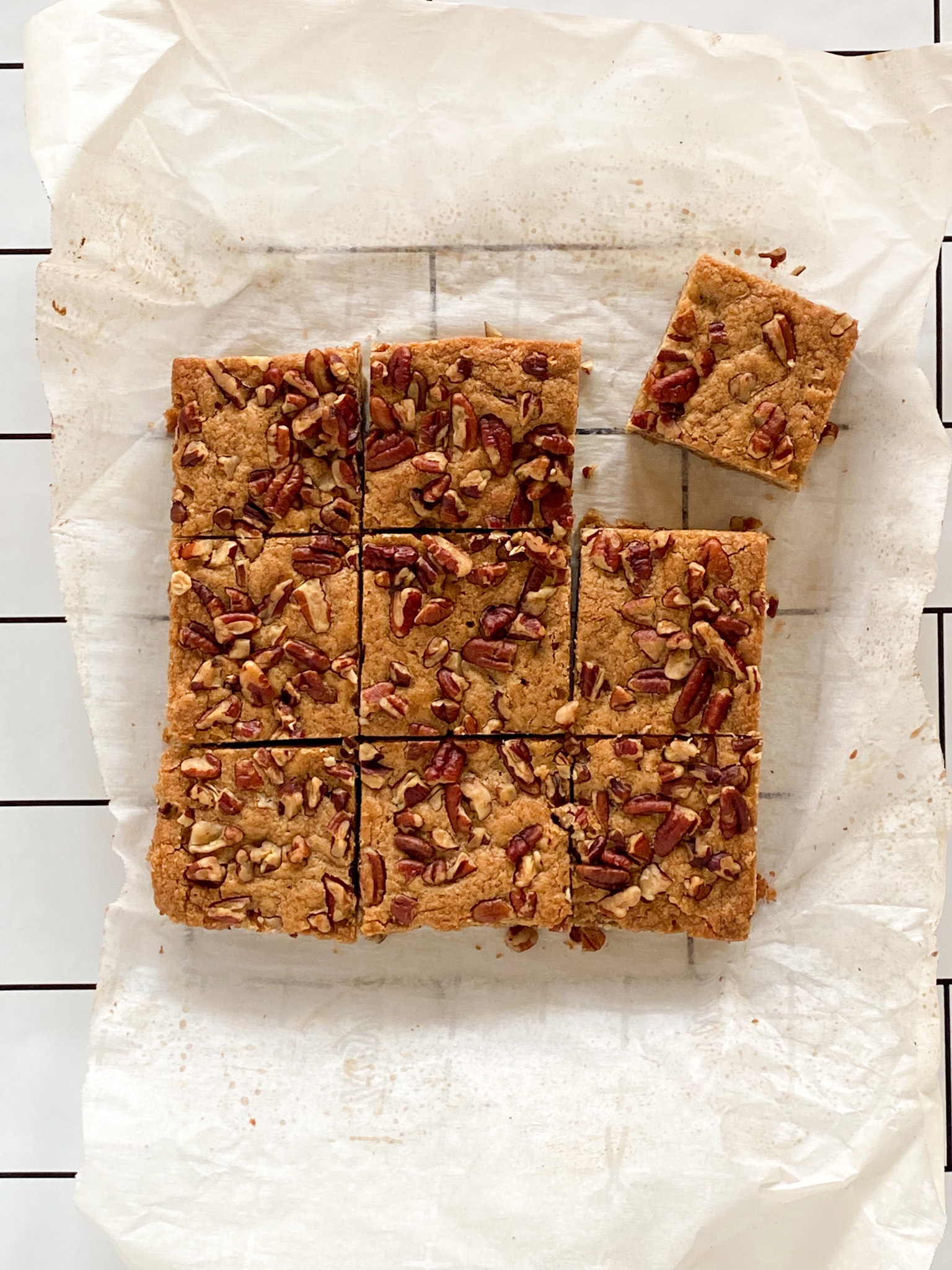 Pecan-covered blondies sit on a piece of parchment paper atop a white tiled surface.