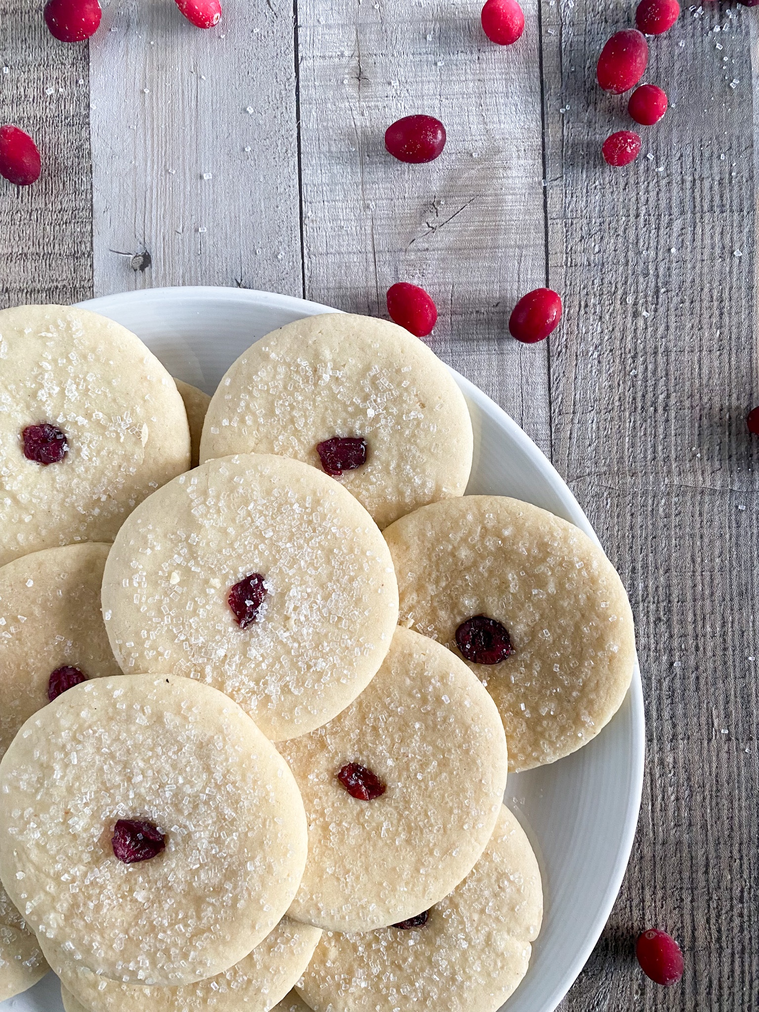 round sugar cookies with a cranberry in the center sit on a white plate on a wooden surface.