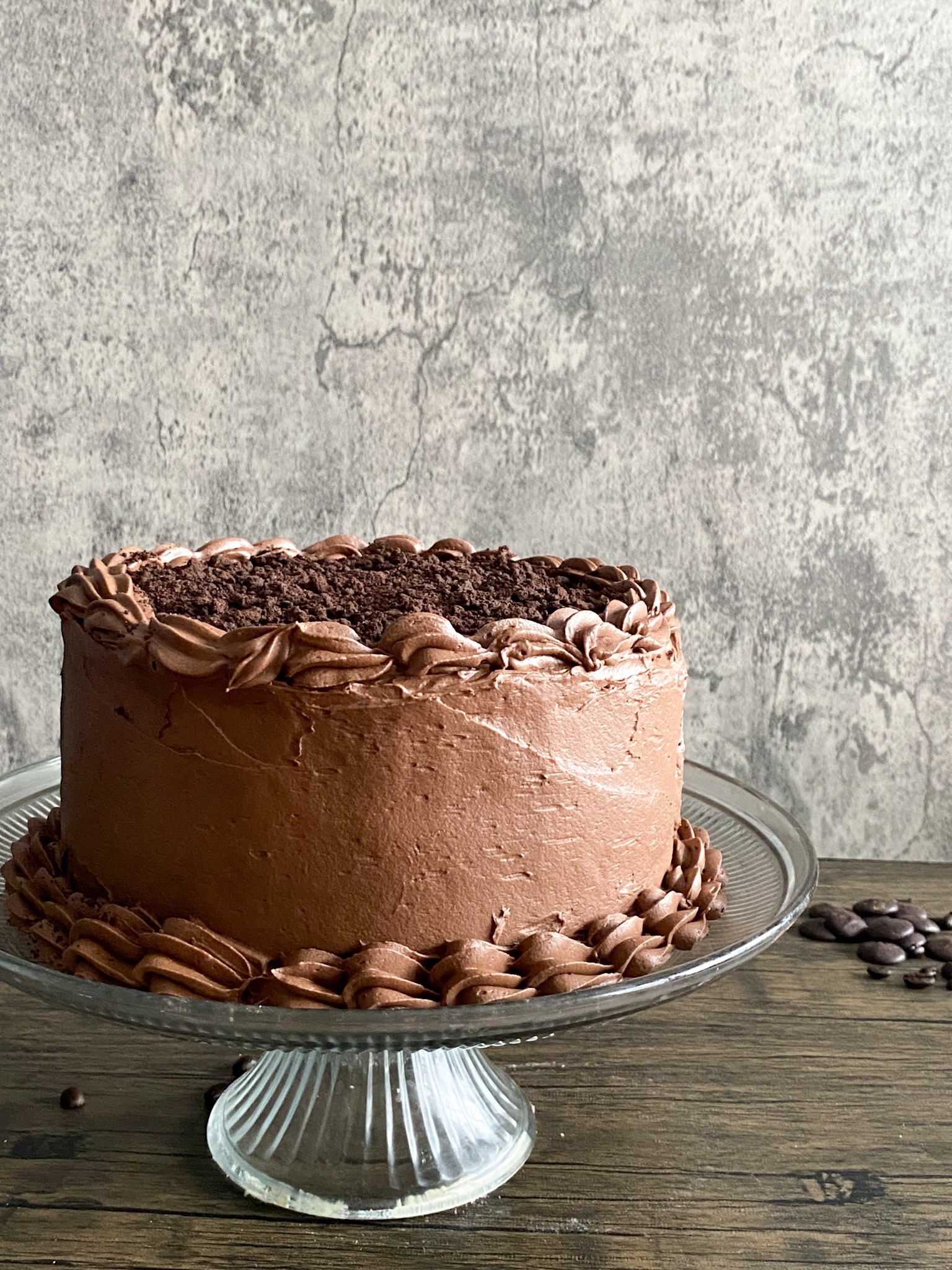 a chocolate layer cake sits on a glass cake stand on a wooden surface with a speckledgrey background