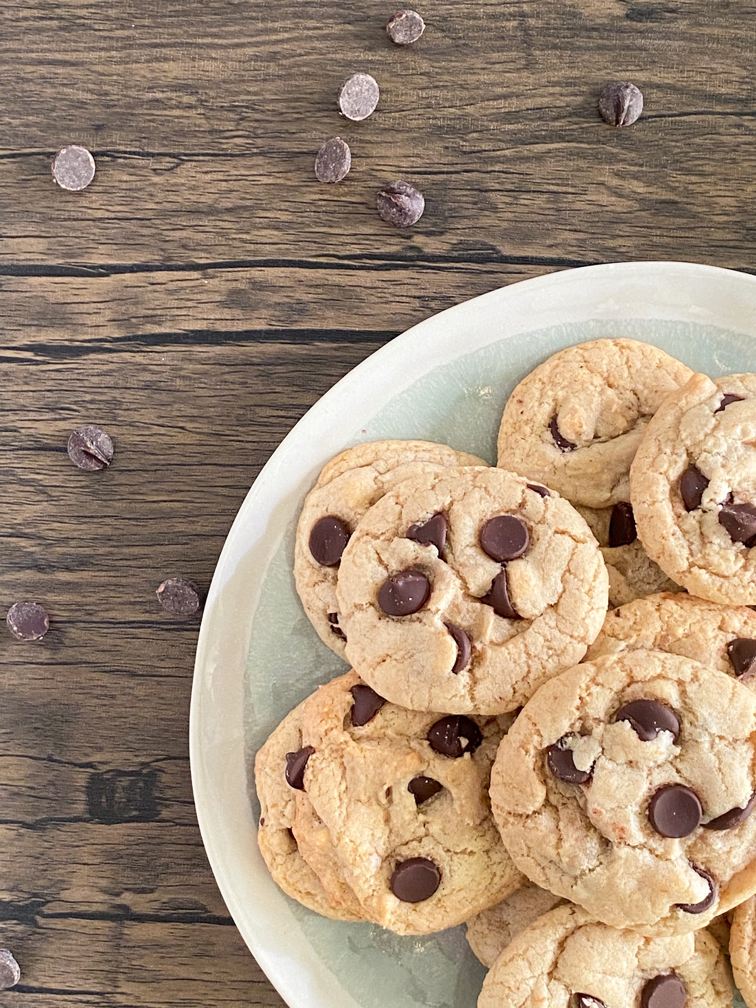 soft vegan chocolate chip cookies are piled on a blue and grey plate that sits on dark wood grain.
