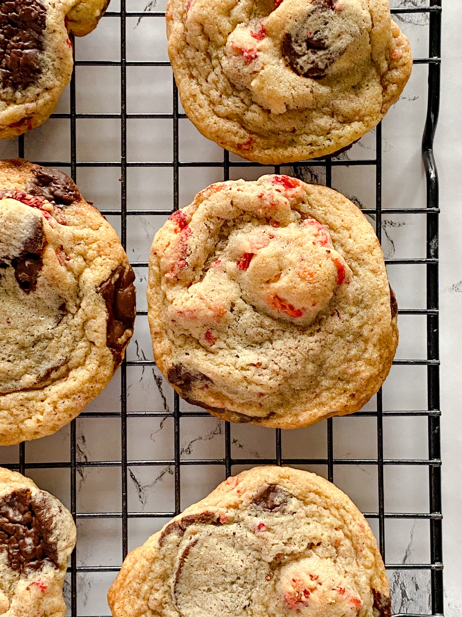 Chocolate chip cookies with strawberries mixed in sit on a cooling rack on a marble-looking surface.