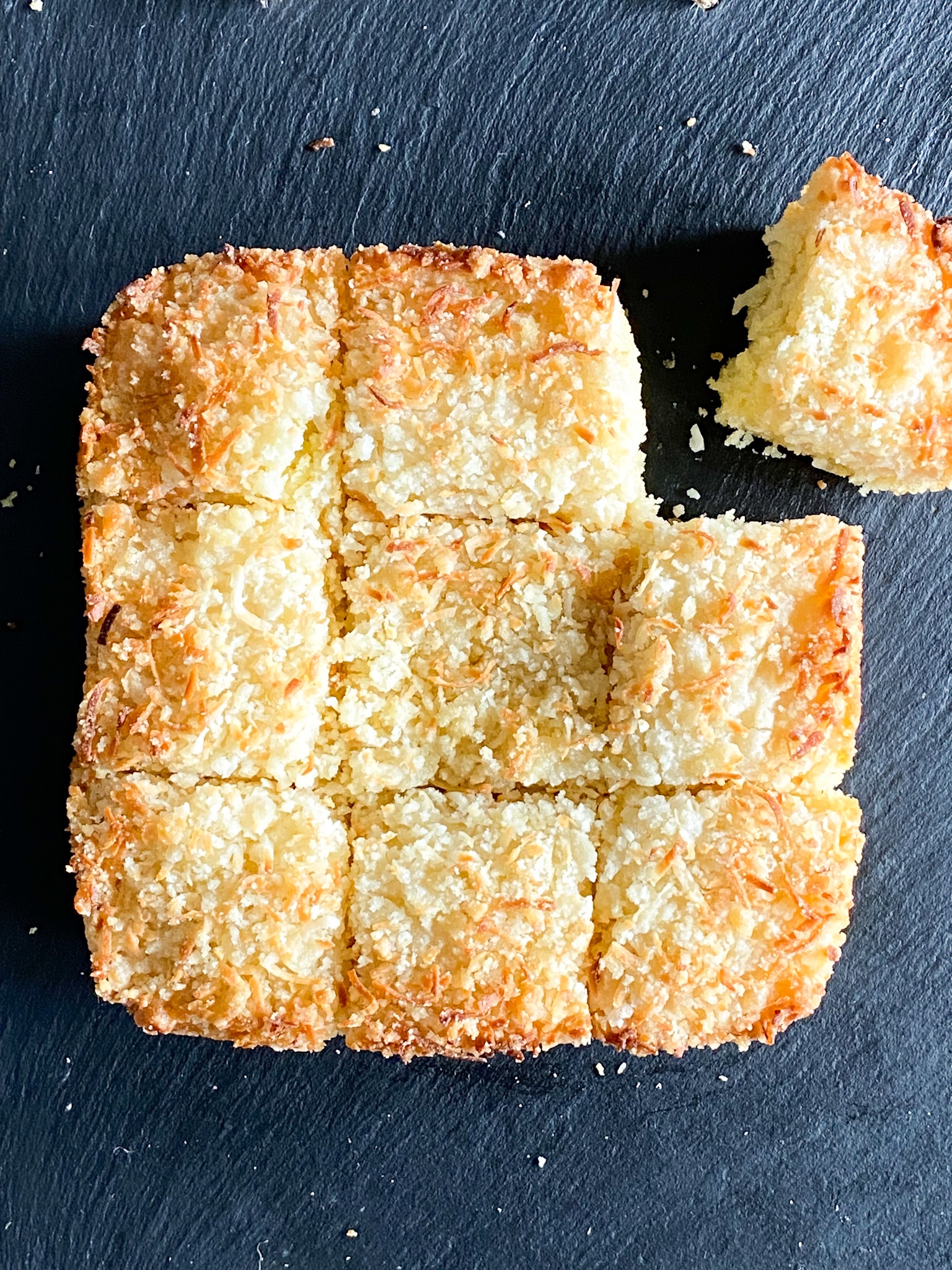 A square cake topped with toasted coconut and cut into nine pieces arranged on a slate surface.