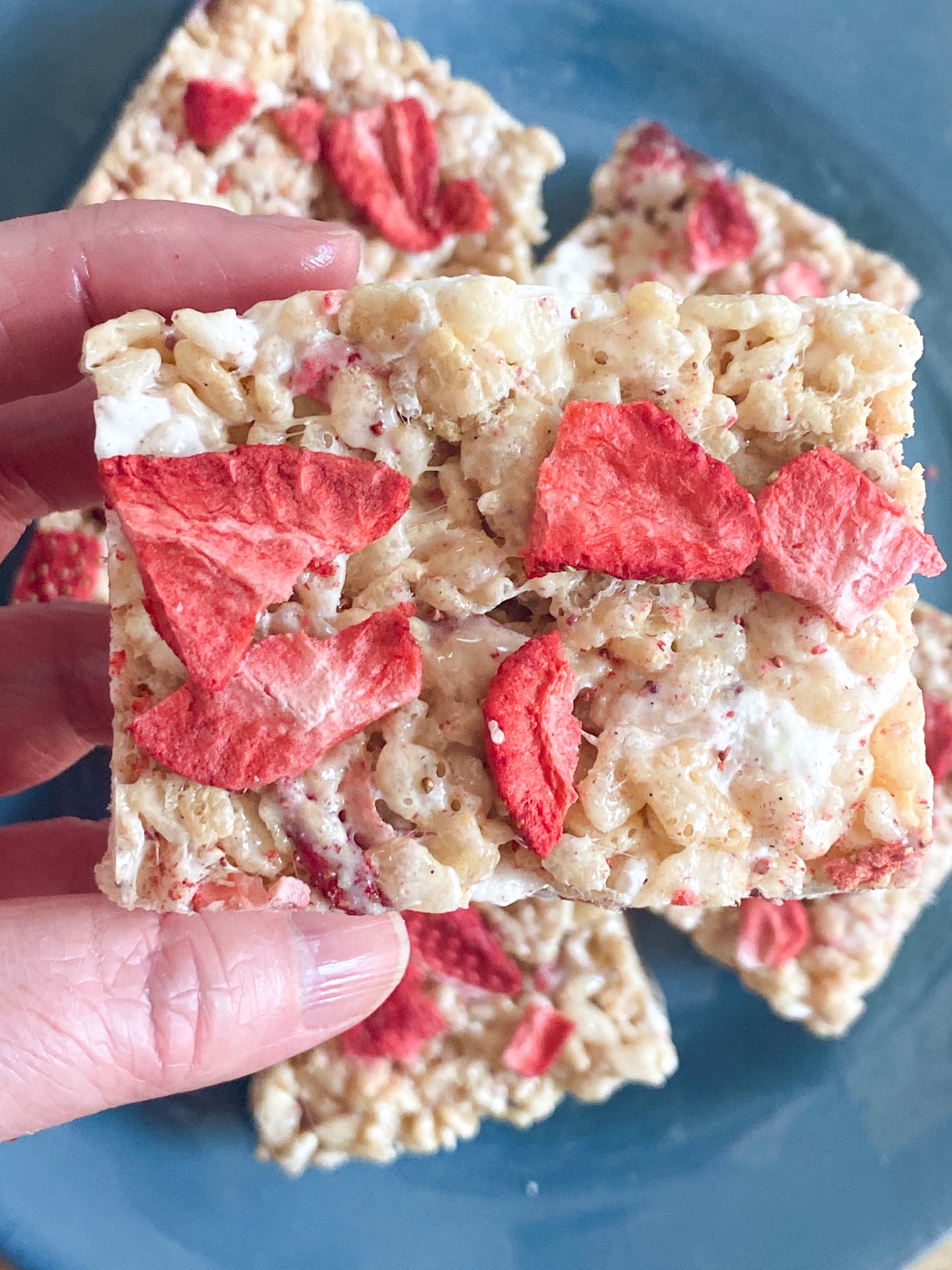 A rice krispie bar topped with freeze-dried strawberries held over a blue plate.