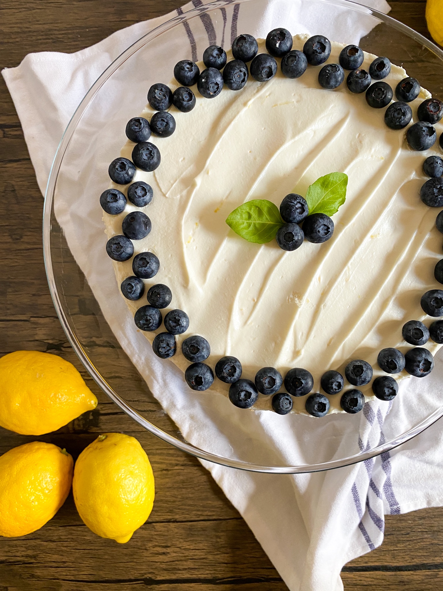 a lemon no-bake cheesecake topped with blueberries sits on a glass plate next to lemons on wood.