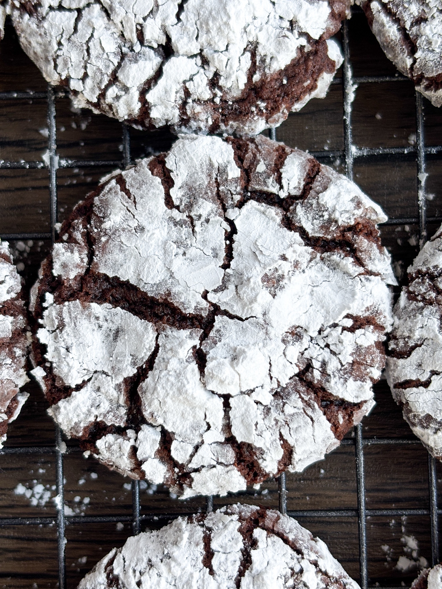 Chocolate crinkle cookies covered in powdered sugar sit on a black baking rack.
